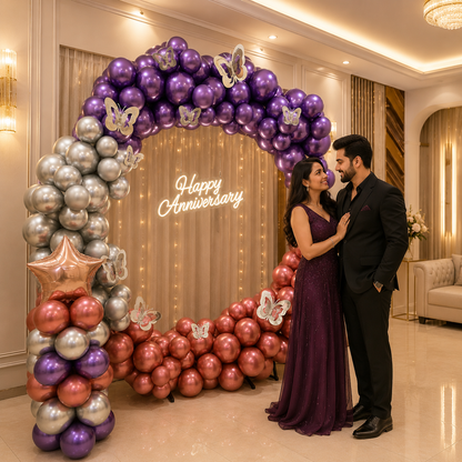 Couple standing in front of a 'Happy Anniversary' balloon arch in a decorated room.