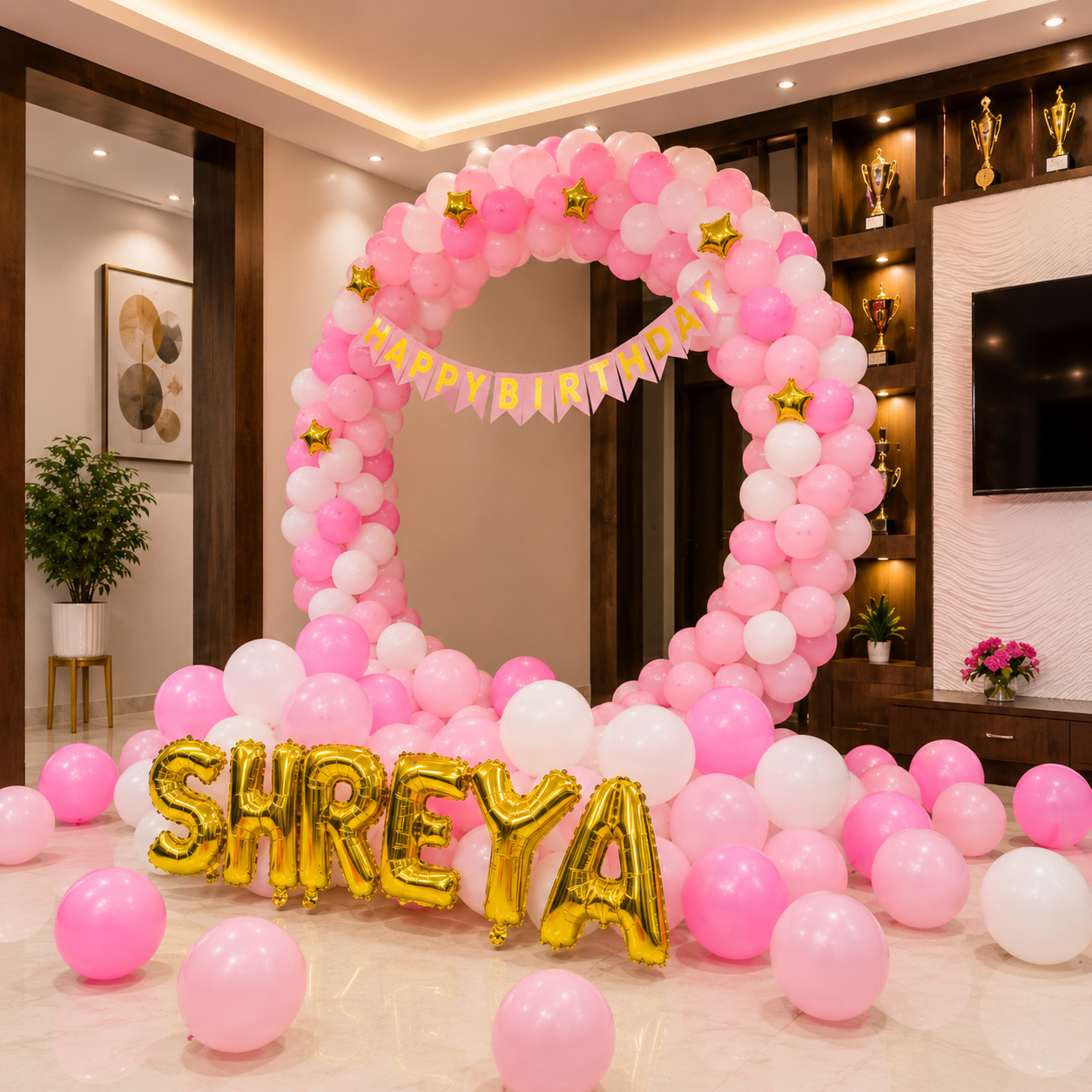 Pink and white balloon arch with 'Happy Birthday' banner and gold 'SHREYA' letters in a room.