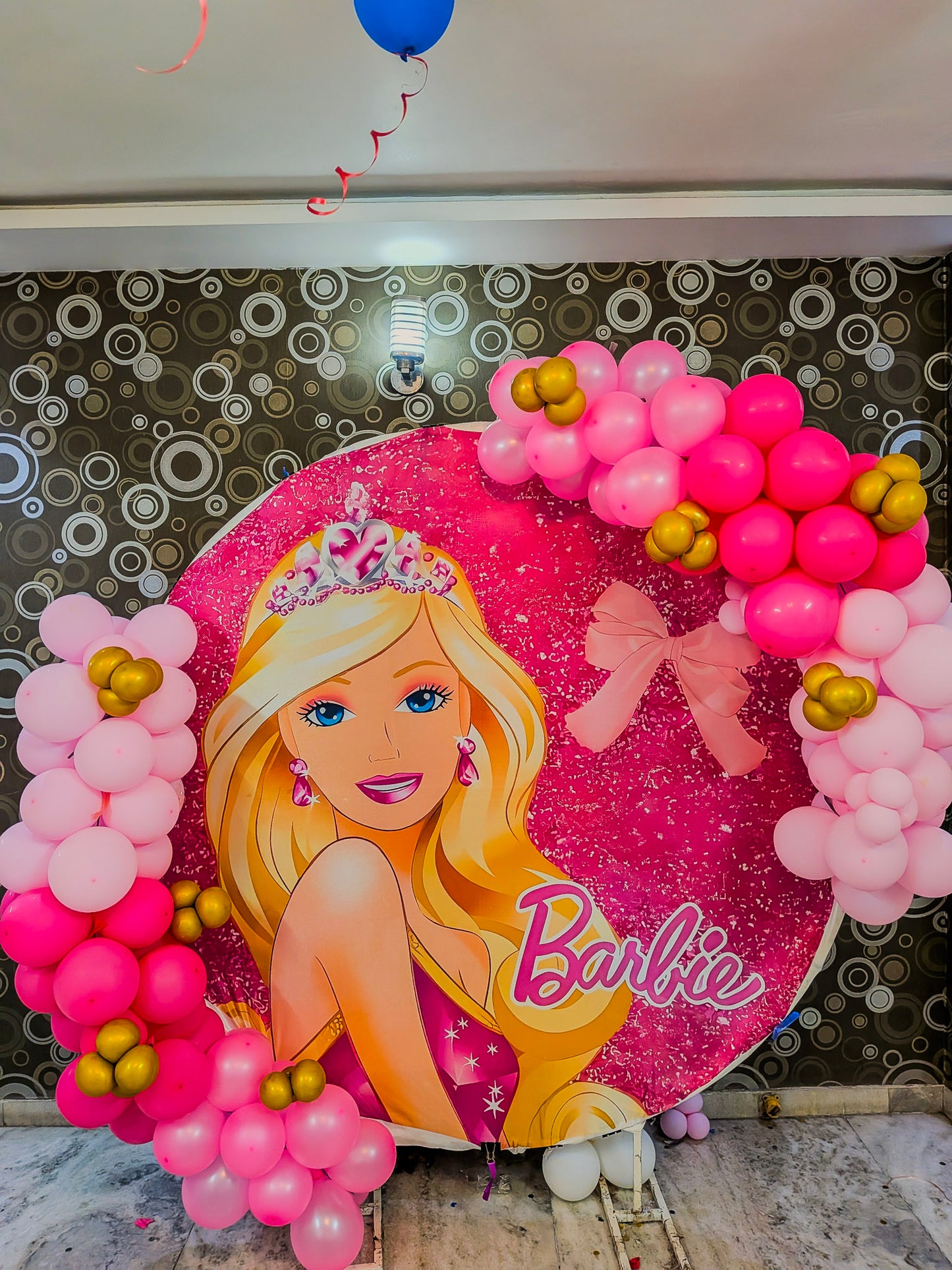 Decorative sign with Barbie design surrounded by pink and gold balloons on a patterned wall.