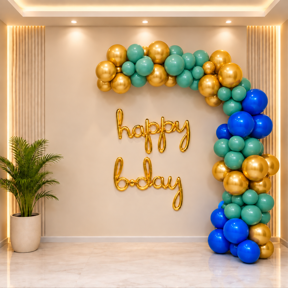 A decorative party backdrop with a balloon garland arch in blue, teal, and gold, plus gold 'happy birthday' lettering on a beige wall in a modern indoor setting.
