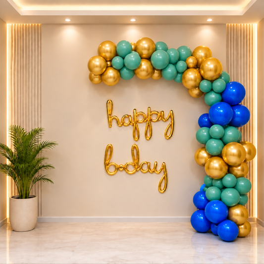 A decorative party backdrop with a balloon garland arch in blue, teal, and gold, plus gold 'happy birthday' lettering on a beige wall in a modern indoor setting.