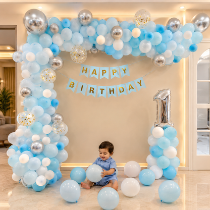 Child under a blue and silver balloon arch with 'Happy Birthday' banner and number '1'.