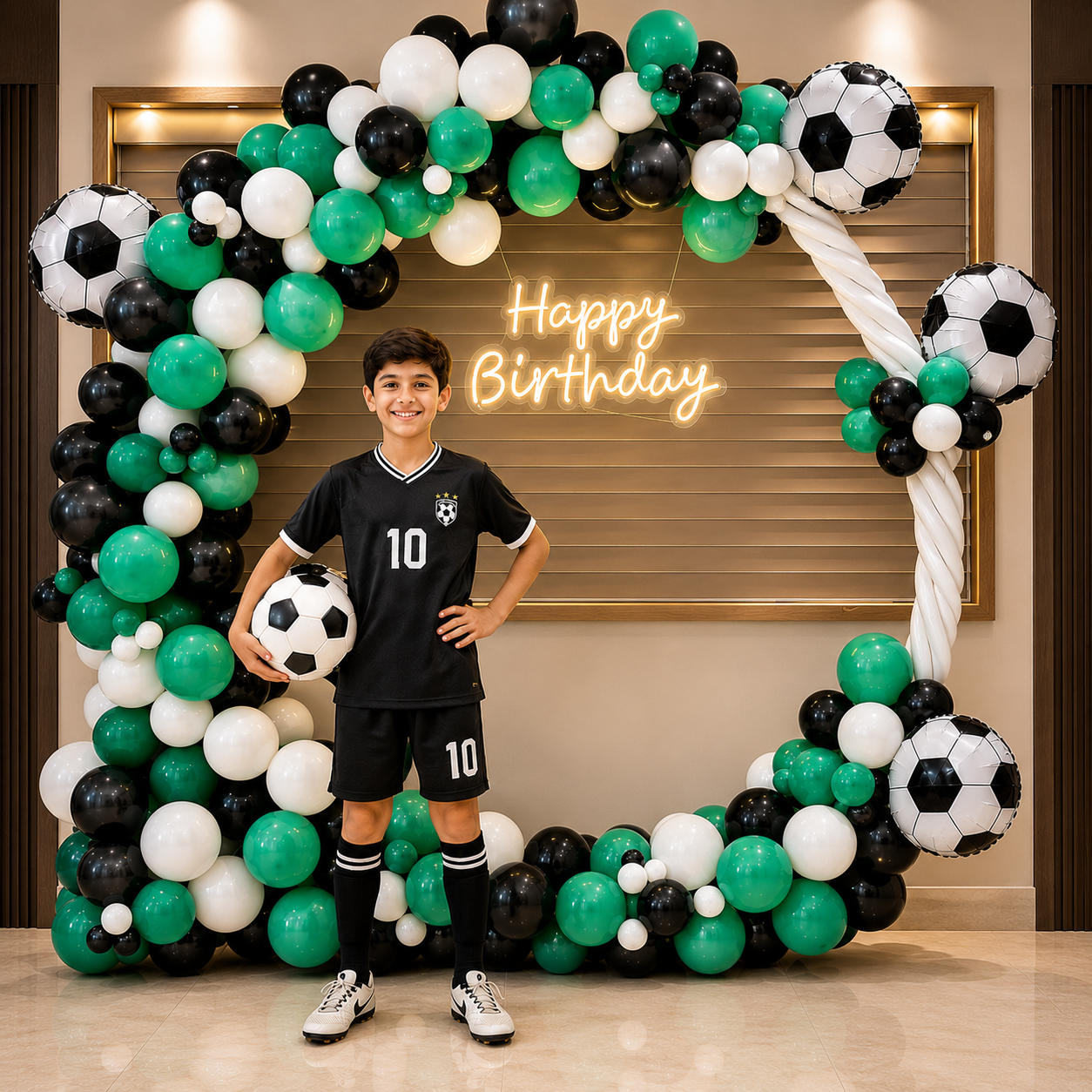 Boy in sports-themed outfit standing in front of a 'Happy Birthday' balloon arch with soccer ball decorations.