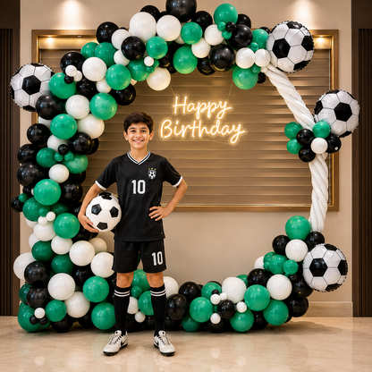 Boy in sports-themed outfit standing in front of a 'Happy Birthday' balloon arch with soccer ball decorations.