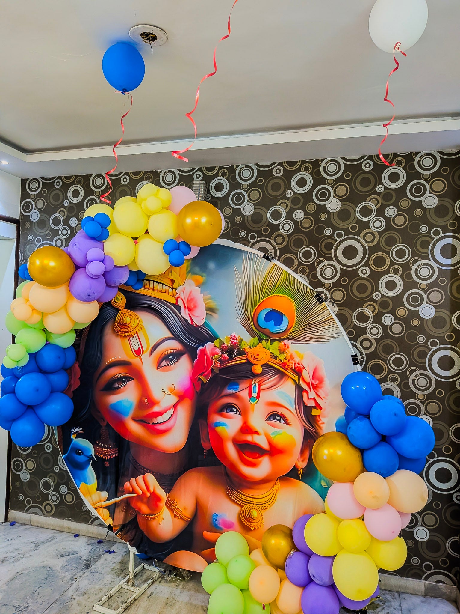 Decorative backdrop with colorful balloons and a portrait of a woman and child Krishna theme with face paint against a patterned wall.
