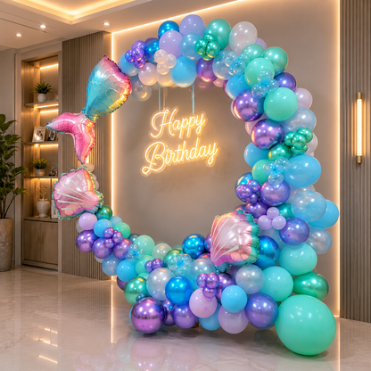 Colorful balloon arch with mermaid tail balloons and 'Happy Birthday' neon sign in a room.