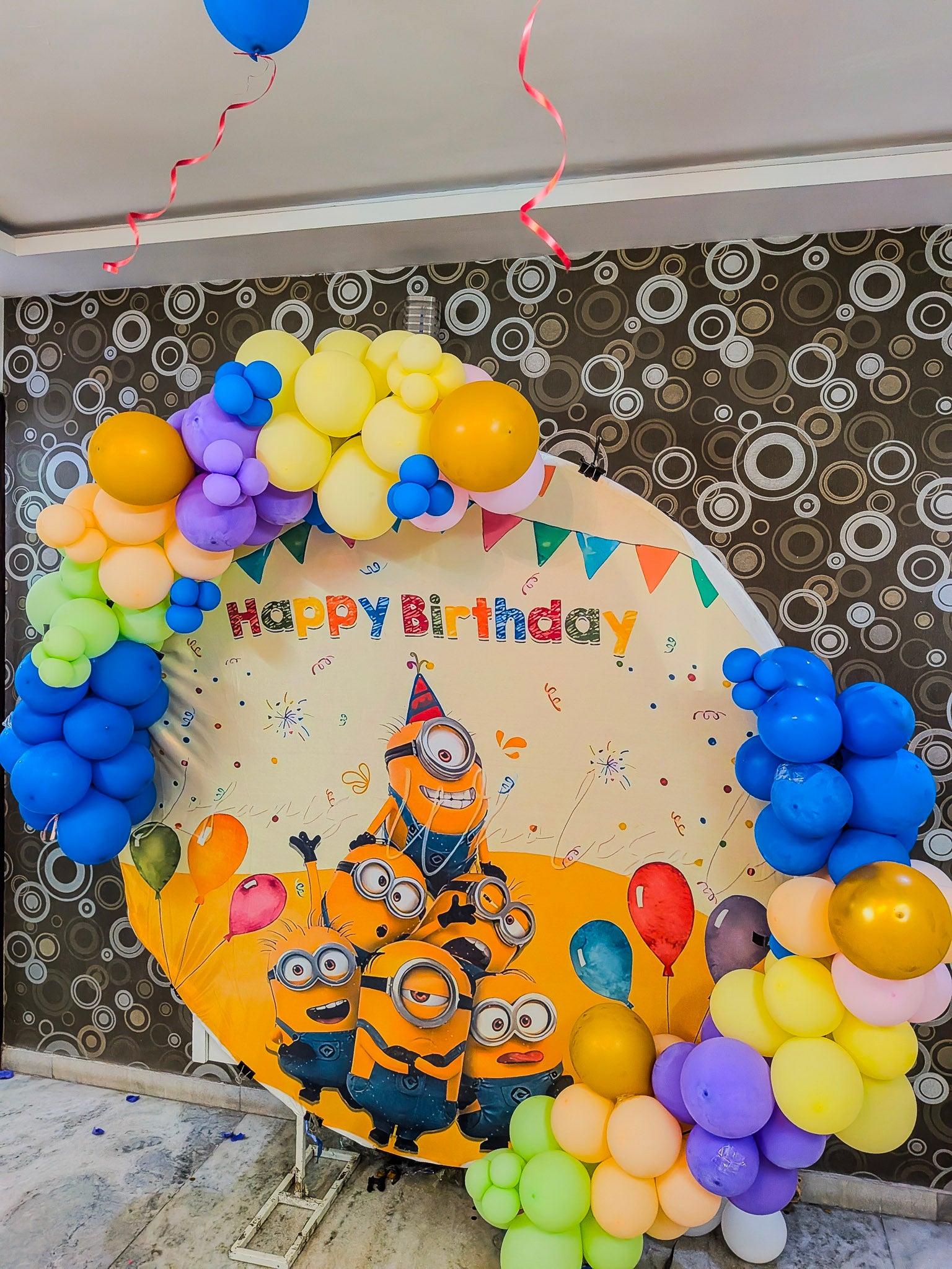 Decorative balloon arch with 'Happy Birthday' sign and Minions design against a patterned wall.