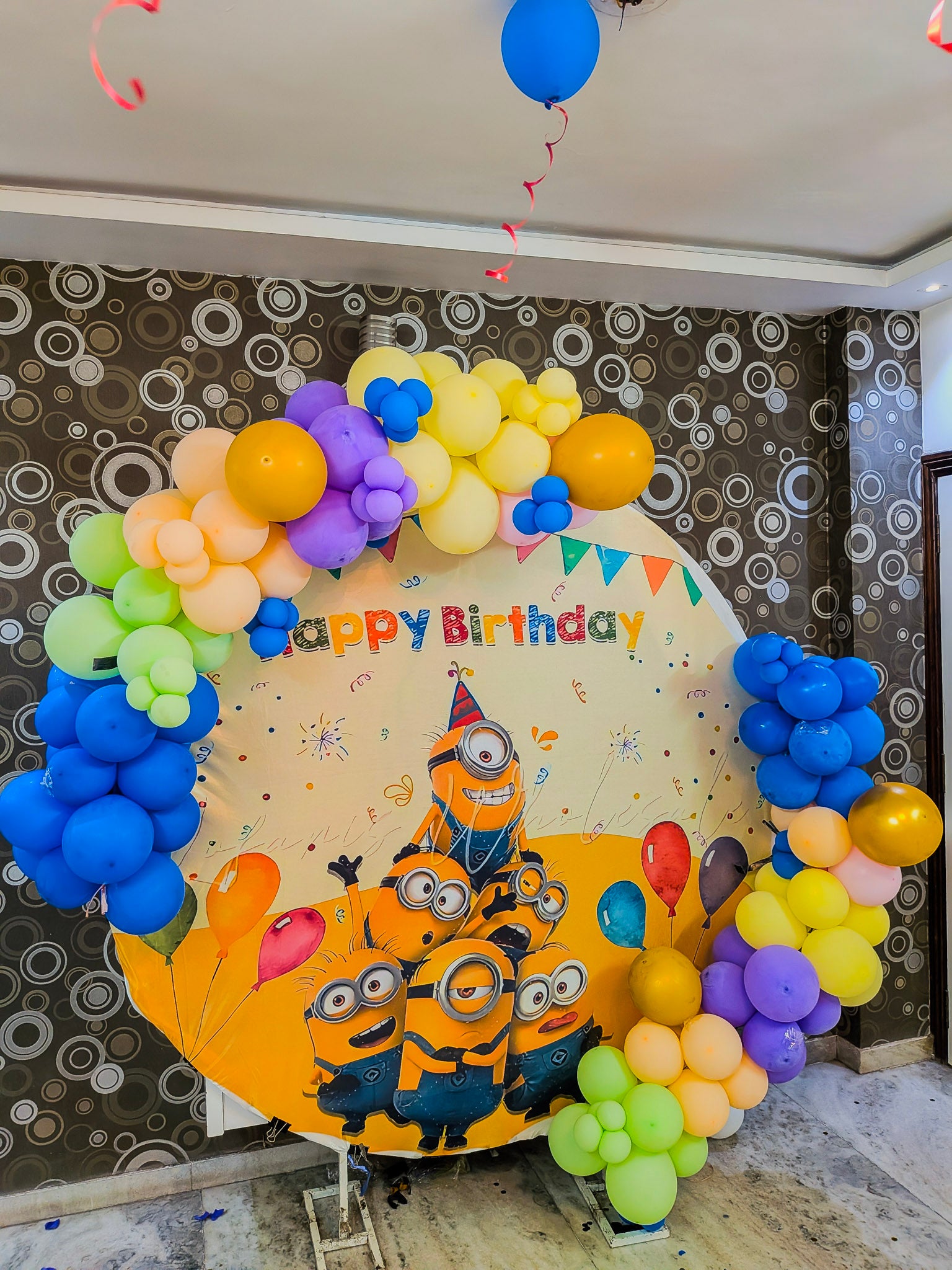 Colorful balloon arch with 'Happy Birthday' sign featuring Minions design on a patterned wall.