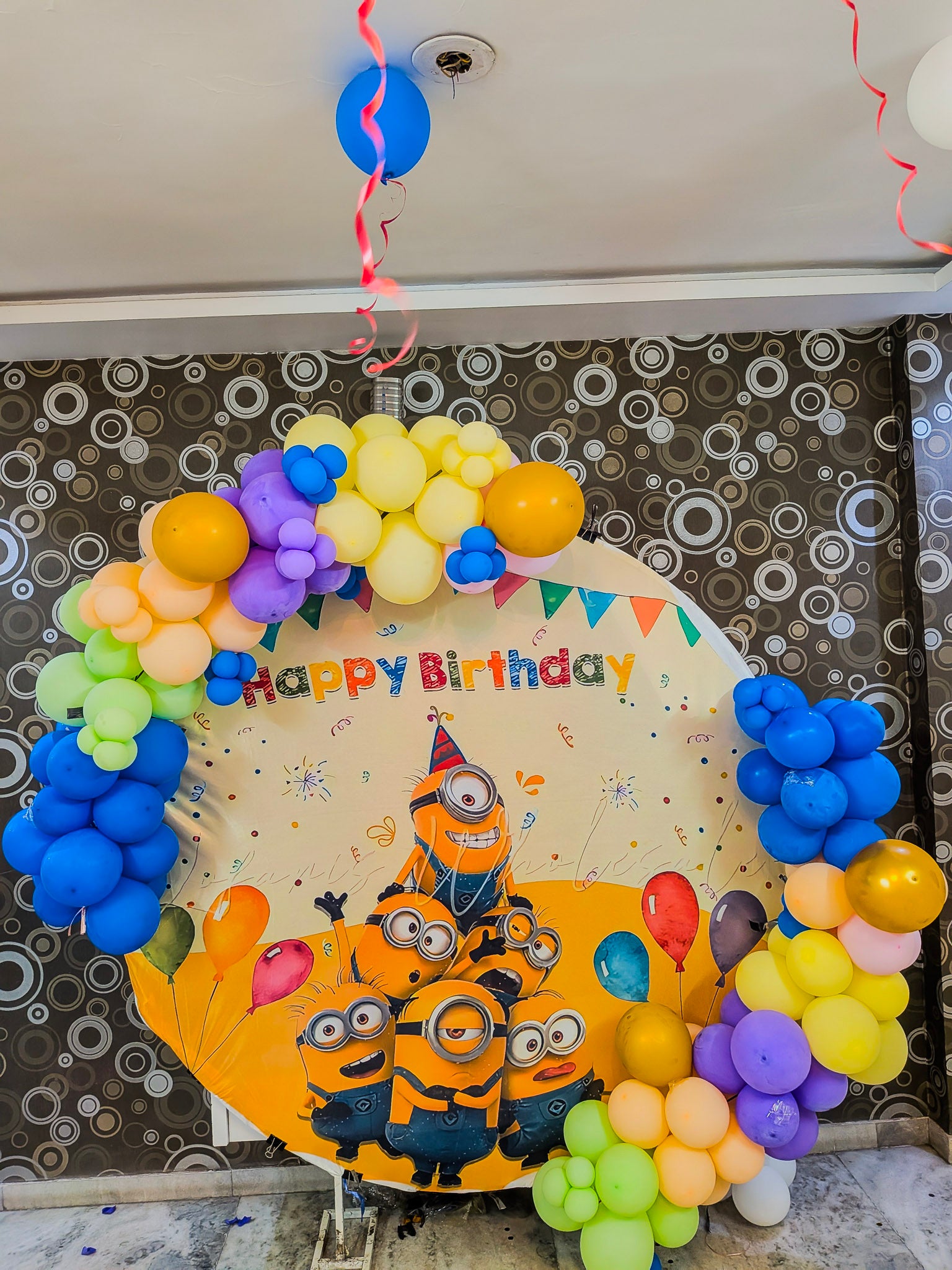 Birthday balloon arch with Minions design and 'Happy Birthday' text on a patterned wall.