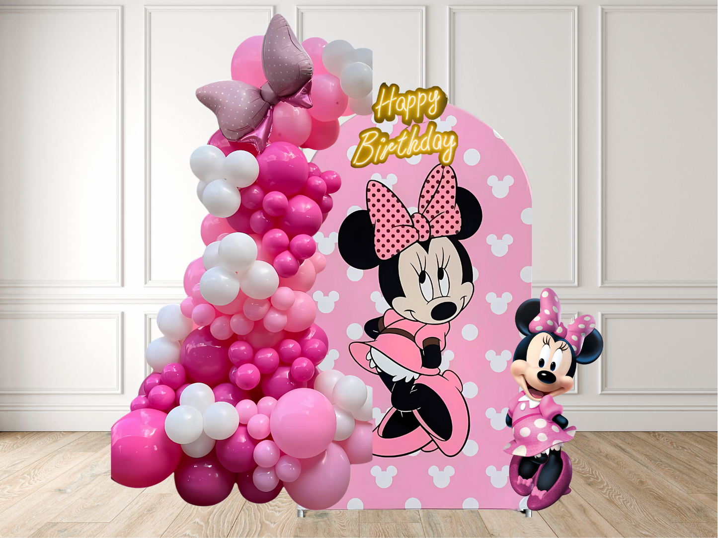 Minnie Mouse-themed birthday balloon arch with a small Minnie Mouse figure on a wooden floor.