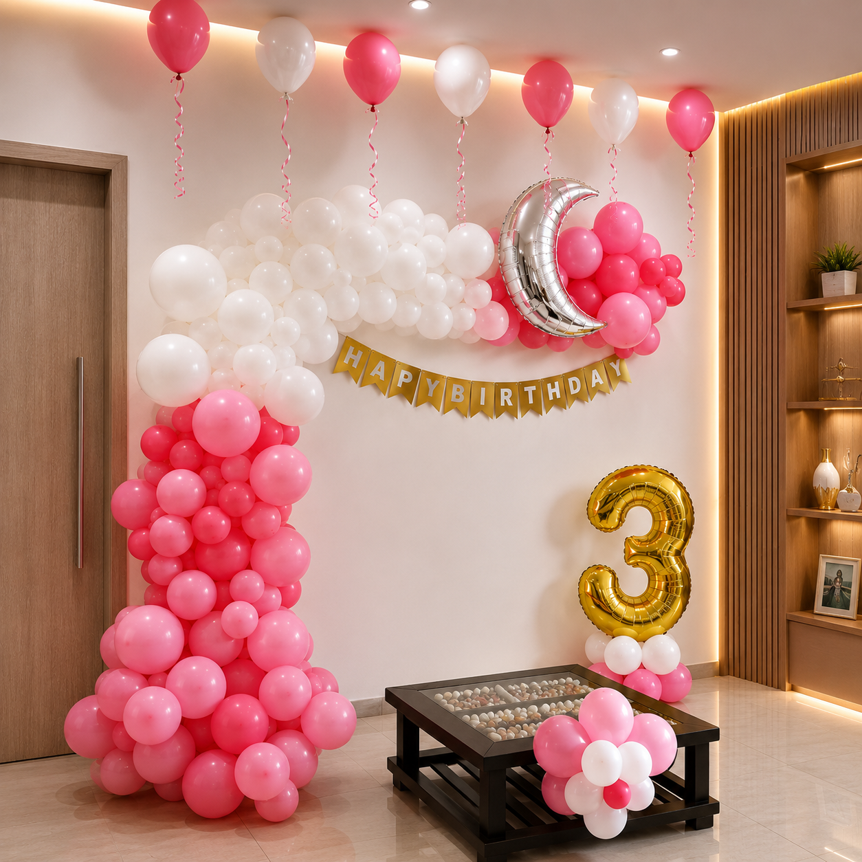 Birthday party setup with pink and white balloons, a 'Happy Birthday' banner, and number '3' balloon in a room.