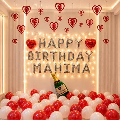 Birthday decoration with 'Happy Birthday Mahima' balloons, red and white balloons, and a bottle of champagne.